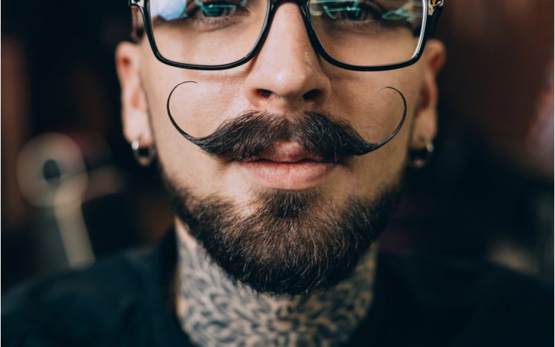 Styled Verdi Type of Beard on a hipster man in dark glasses, a black shirt, and with neck tattoos