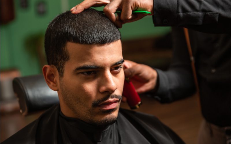 #5 Buzz Cut With Straight-Across Fringe, a popular haircut for men, on a guy in a barber chair with a mustache