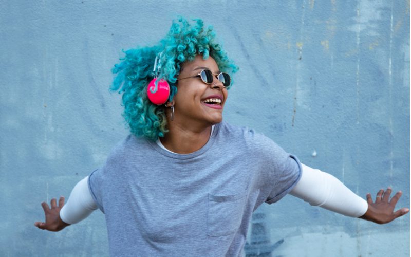 Aquamarine Blue hair color on a brown skin woman wearing headphones and spreading her arms against a blue wall