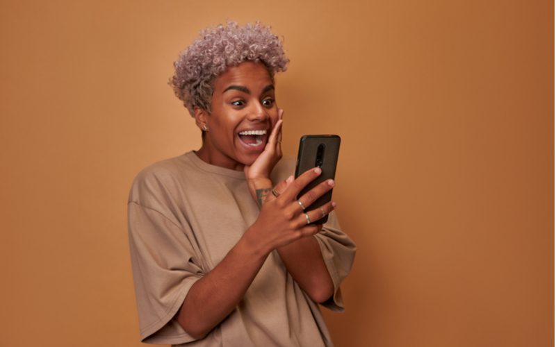 Violet-Toned Silver hair color on a brown skinned woman holding her left cheek in excitement after seeing something she liked on her smartphone