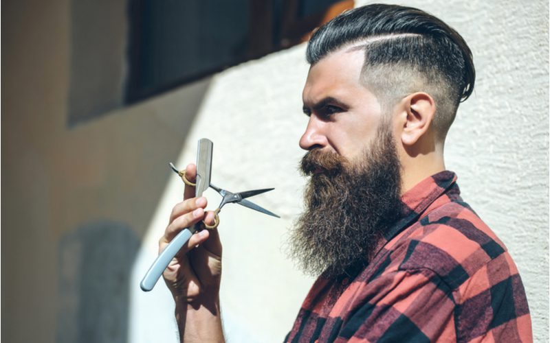 Long Comb Over Skin Fade With Hard Part on a lumberjack-looking guy holding beard trimming scissors