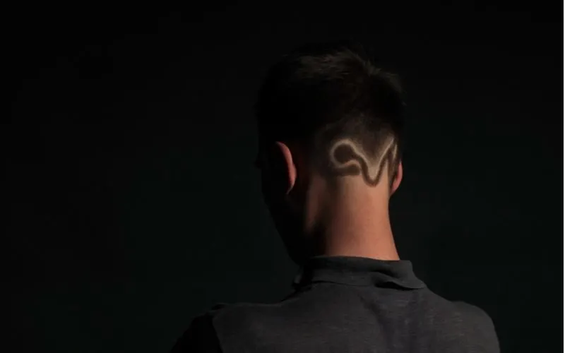 Guy in a dark room looking away from the camera with a hairstyle pattern shaved into the back of his head