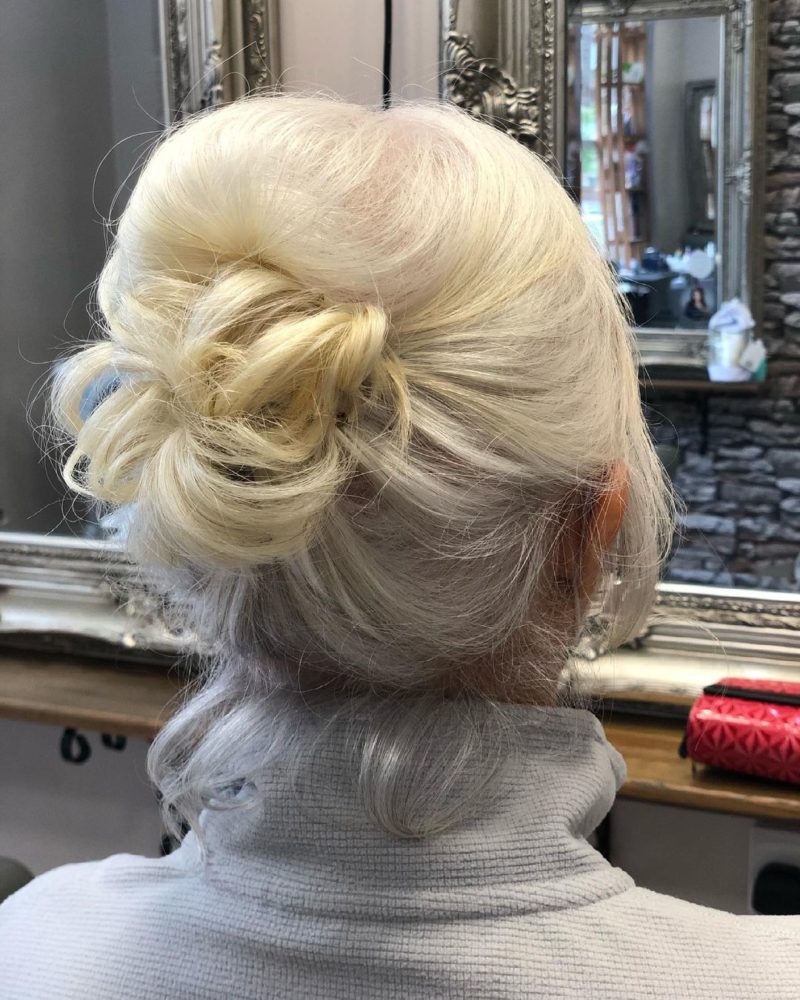 Fancy Updo on the hair of a blonde woman in a white turtleneck listed as an idea for a wedding guest hairstyle