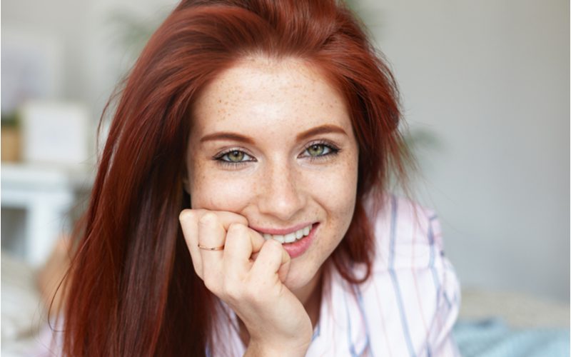 Woman with auburn hair leaning on her right palm and smiling while gazing at the reader from the top of her eyes