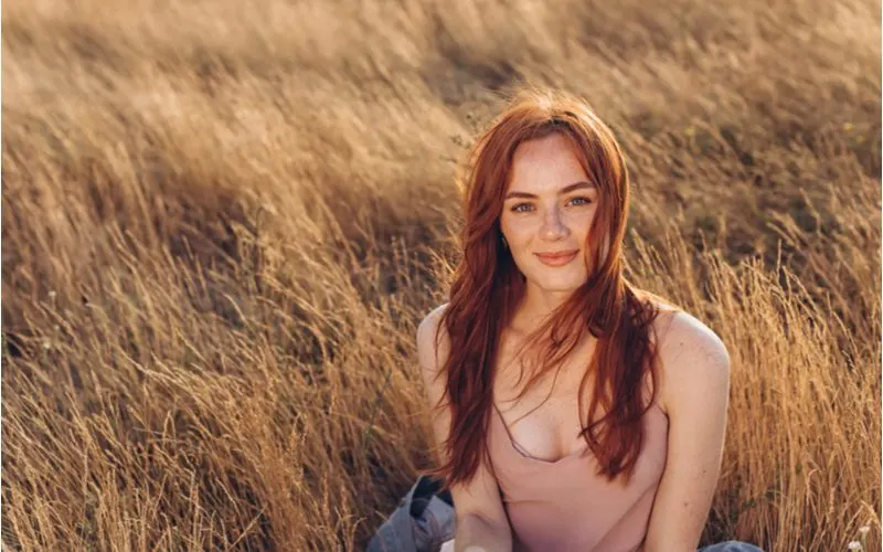 Lady wearing a light pink camisole with a ton of cleavage exposed while also rocking auburn hair while sitting in a wheat field