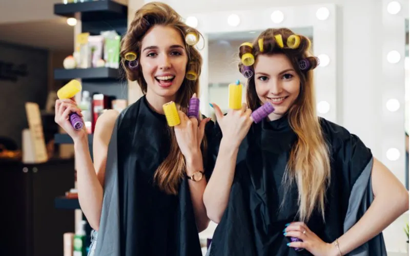 Teen girl hairstyle being done with rollers as the two girls in capes and roller stand next to each other and smile