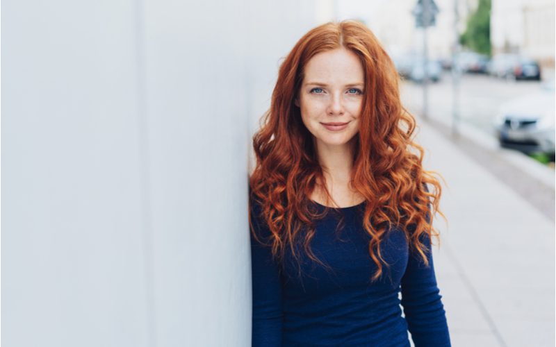 Bright Ginger-Toned Auburn hair on a thin woman in a blue sweater standing against a white walled building