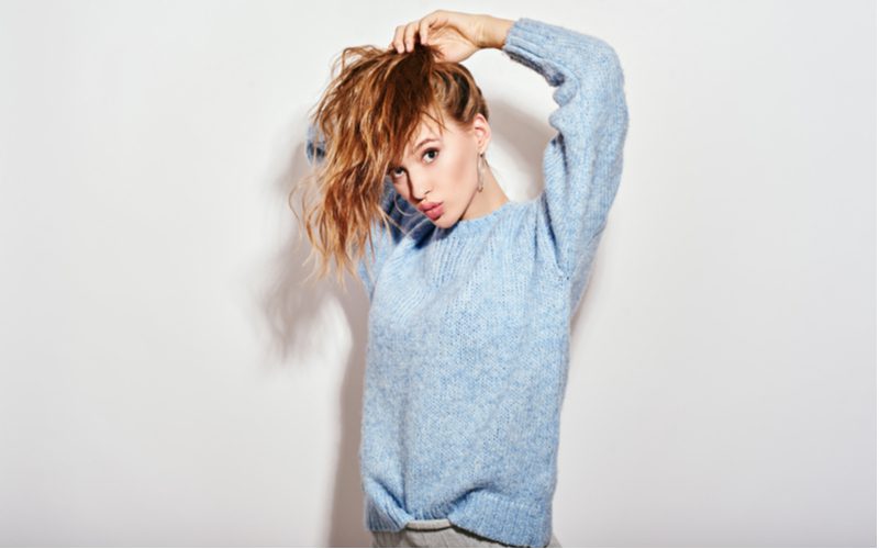 Woman in a blue-gray sweater putting her hair up into an easy hairstyle that will be a messy bun