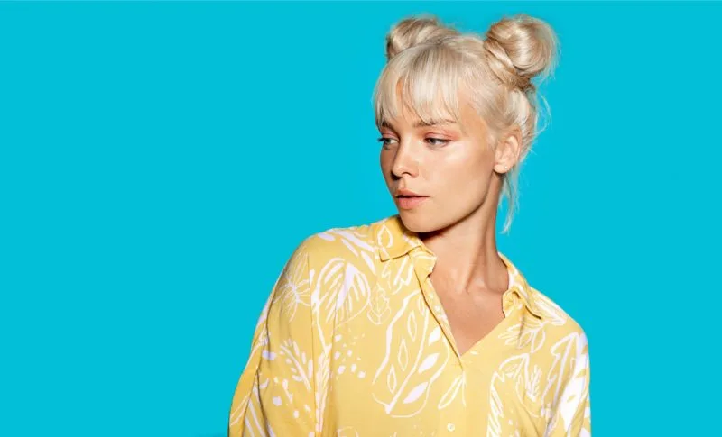 Blonde woman in a blue room and a yellow shirt wearing grown-up space buns, a featured long to medium length hairstyle