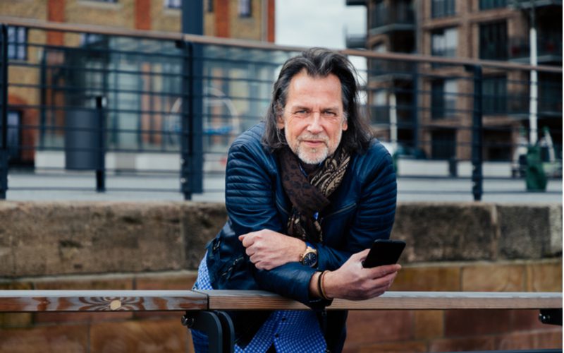 Older man with long hair leans over a metal railing and looks at his phone