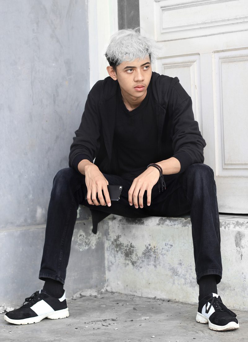 For a roundup of messy hair boys, a guy wearing a Messy Two-Toned Two Block Cut sits on a concrete step