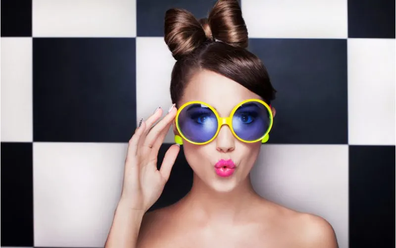 Gal in a black and white checkered room with yellow round glasses holding the right side of the face and making a duck face pose with her lips for a piece on easy hairstyles