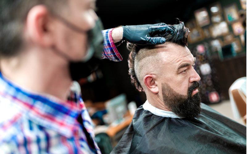 Guy in a barber's chair getting a mohawk haircut from a gloved and masked barber wearing a blue and red plaid shirt