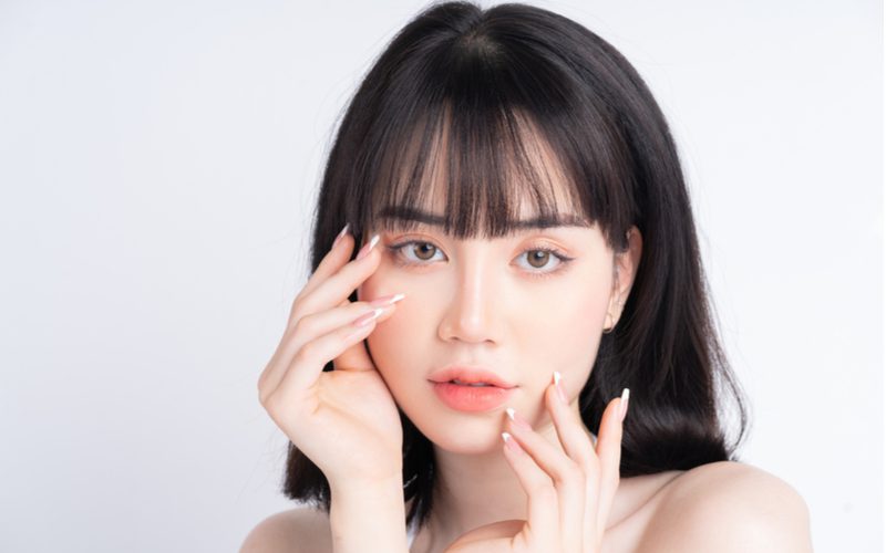 Korean Air bangs, a popular type of bangs, on a fair skinned woman holding her hands to her face in a sensual pose