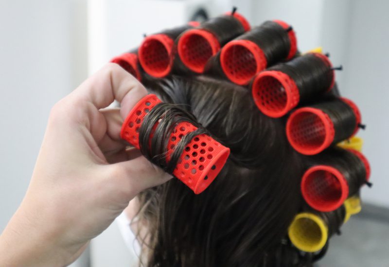 For a piece on heatless curls, a woman with a bunch of plastic heatless curlers in her hair with a hand touching one of them on the left side
