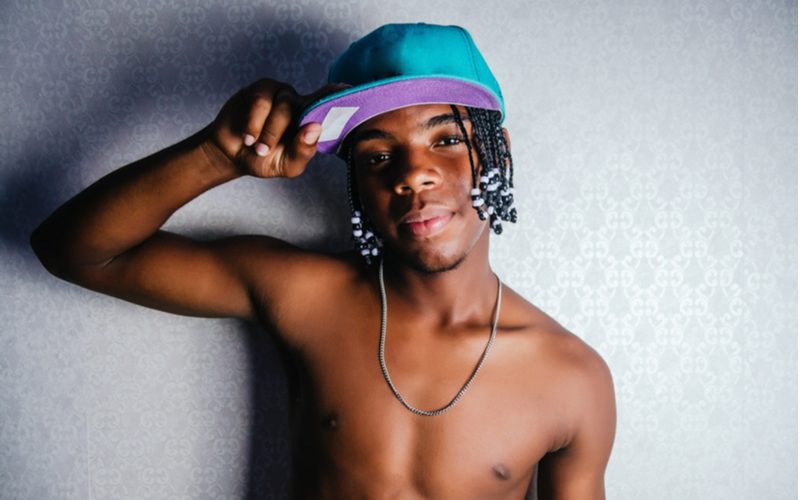 Black male hairstyles featuring a guy with box braids with teal ends wearing a blue hat while holding the brim