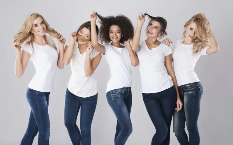 Women of different races and skin colors with all different types of hair from our hair type chart stand in a pink room with dark jeans and white shirts