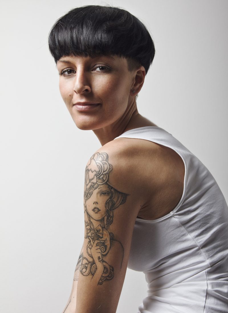 Stacked Undercut Mushroom Haircut on a woman in a white sleeveless shirt in a studio slouching forward with a grin on her face