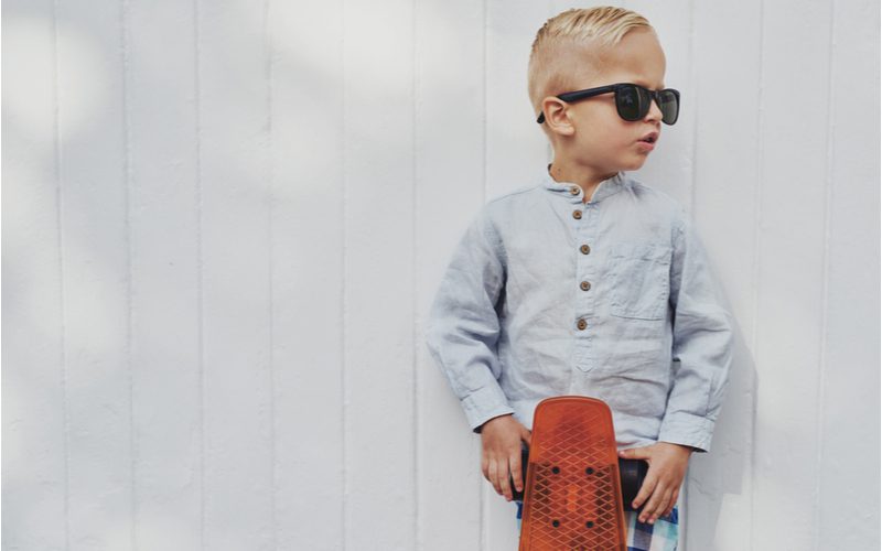 Little boy in sunglasses and a grey button-up shirt holds a longboard