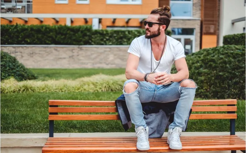 Guy with a man bun and a thick beard sits on the back of a bench and looks to his right while clasping his hands together