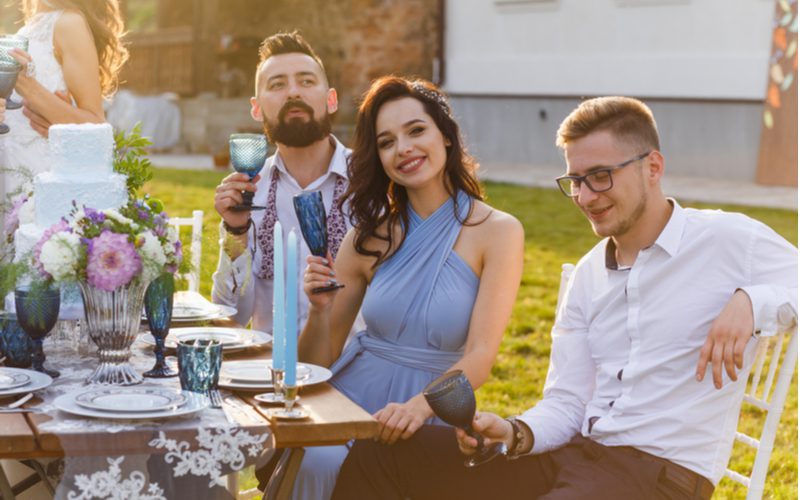 Image for a piece titled wedding guest hairstyles featuring a man and woman sitting at a rustic outdoor table toasting the bride and groom