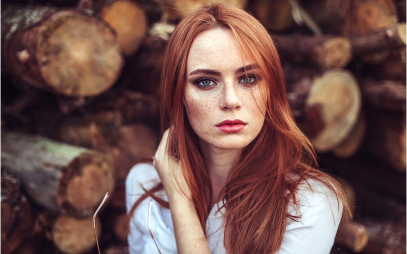 Gilded Auburn hair on a woman with freckles holding the right side of her head in front of a pile of wood