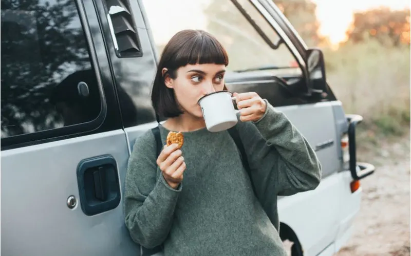 Woman wearing a dark green shirt sips coffee from a mug and leans against a camper van while wearing micro bangs, one of the most popular types of bangs