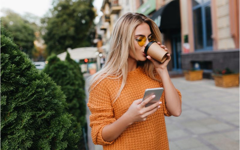 Face-Framing Fringe bangs worn by a woman in an orange sweater checking her phone and drinking a coffee