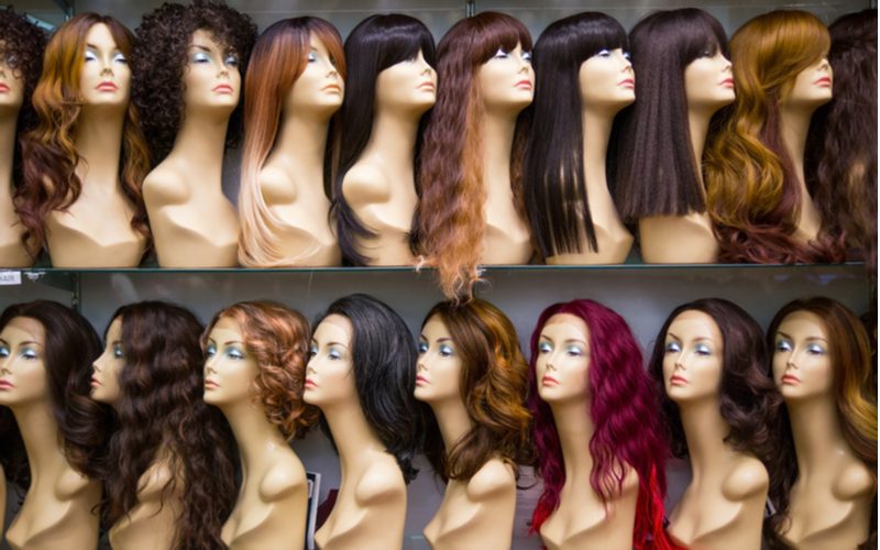 A row of mannequins with various types of wigs on their heads in various colors line the shelves of a store