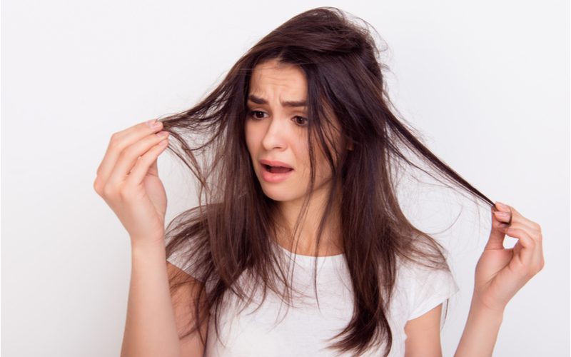 Image for a piece tilted How to Tell If Your Hair Is Damaged featuring a woman in a white shirt looking at the end of her hair in shock because it's damaged