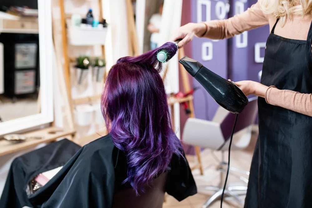 For a piece on dyeing dark hair purple, a woman in a salon with purple hair gets it blow dried