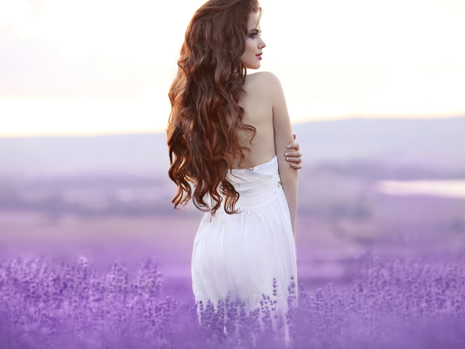 Woman with red waist length hair standing in a white dress in a field of purple flowers