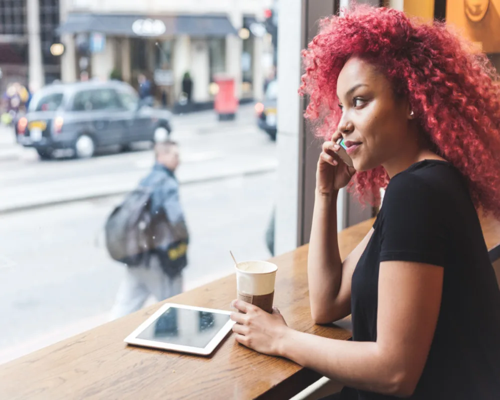 Black woman with red hair in a coffee shop talking on a cell phone