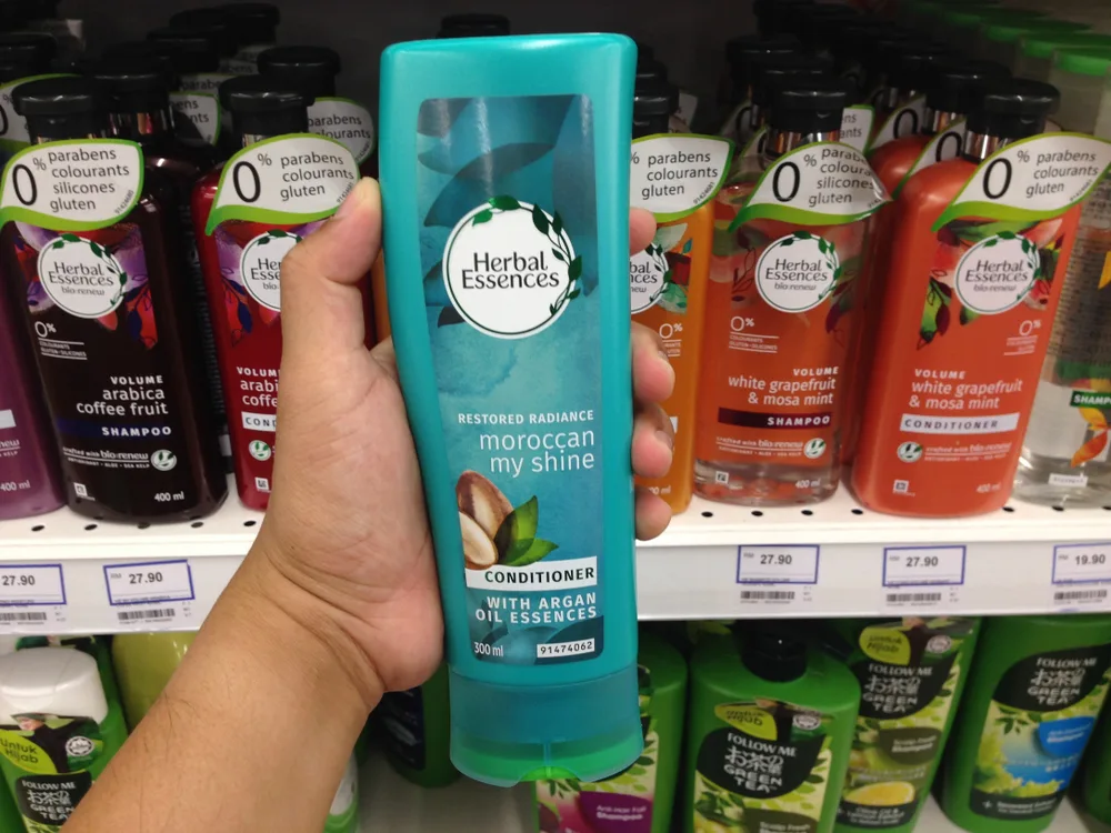 Person holding a shampoo bottle with others in the background for a piece titled Is Herbal Essences Good for Your Hair