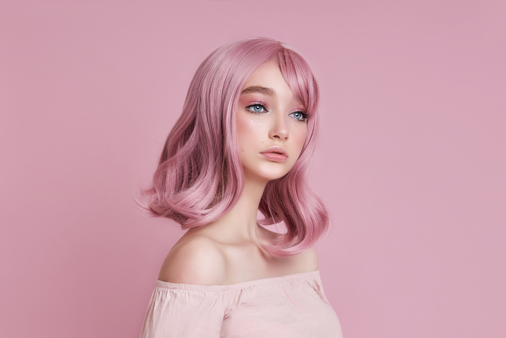Dusty blush pink, a hair color for blue eyes, on a woman in a pink room wearing an over-the-shoulder shirt