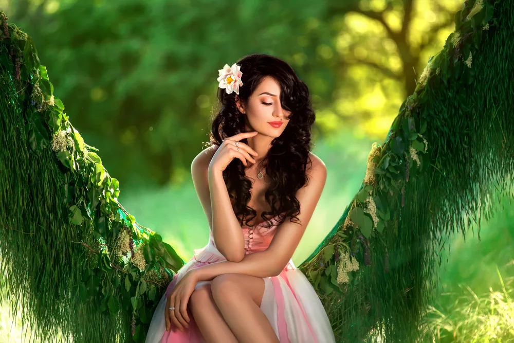 Woman with black hair and light skin wears a flower in her hair and sits on a tree
