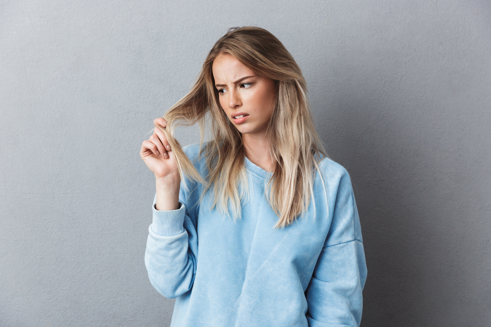 Woman with uneven hair growth looking at one side of her hair in disgust while wearing a blue oversized sweatshirt