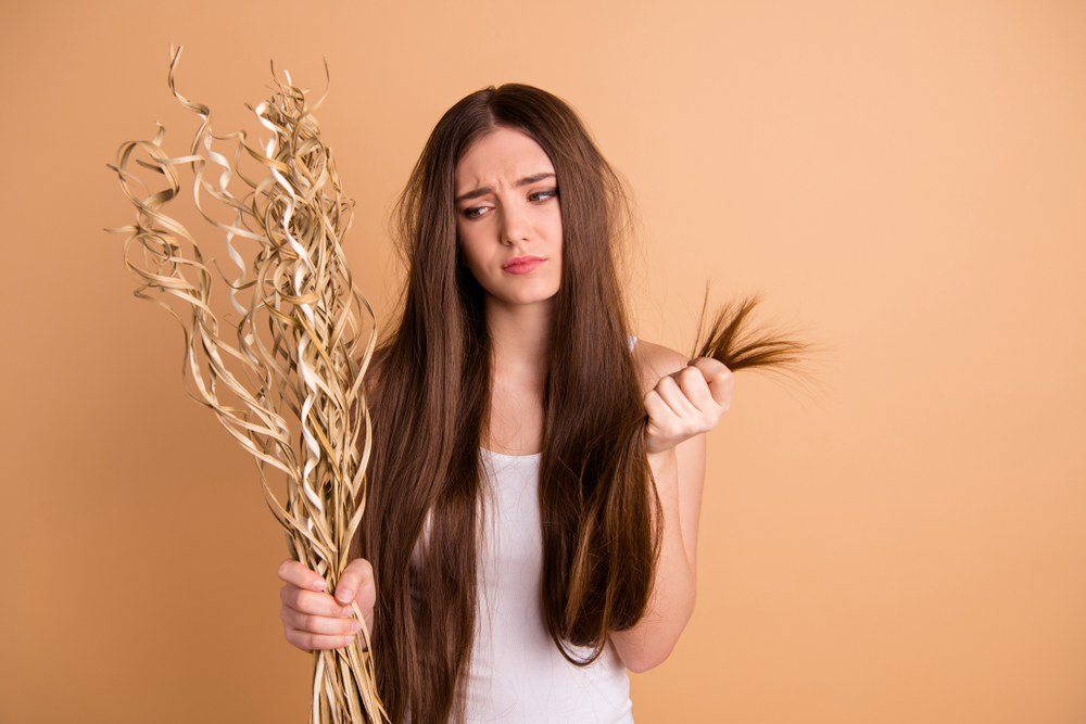 Woman wondering why her hair feels like straw while holding both her hair and a bundle of straw in a brown room