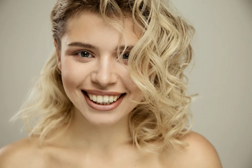 Blonde woman with brown eyebrows smiling at the camera with her shoulders exposed