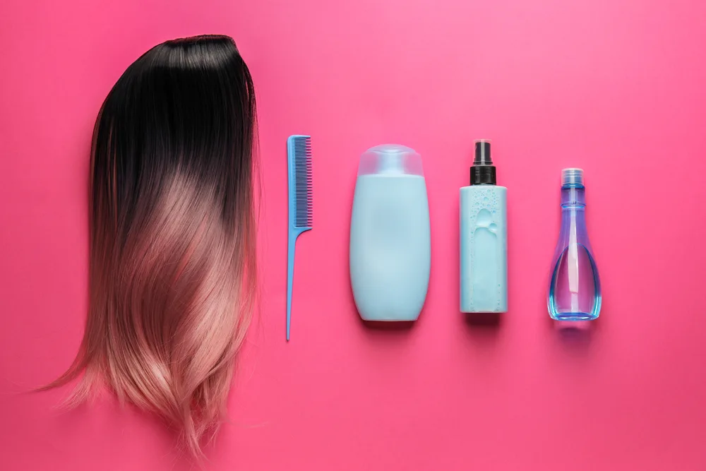 Image for a guide on how to wash a wig with fabric softener with various products next to a wig in a pink layflat image