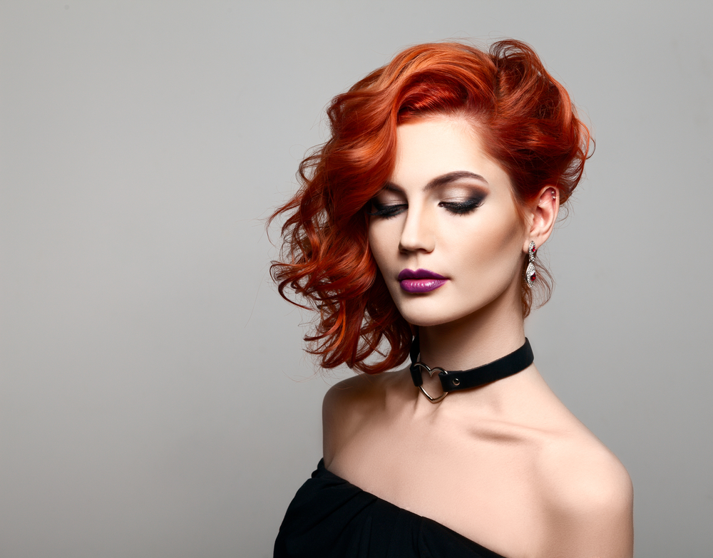 Woman with red hair and pale skin in a black dress stands in a grey room for a piece on can I dye my head red without bleaching