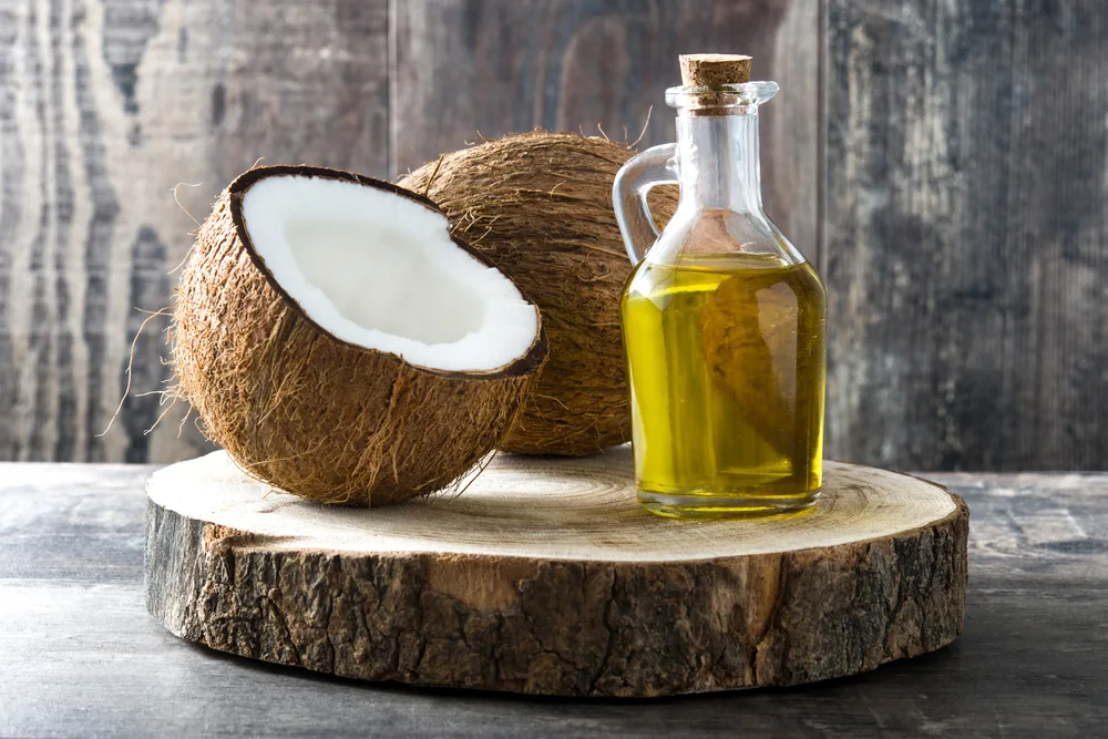 On a wooden table sits a jar of oil and a coconut for a piece titled should i use refined or unrefined coconut oil for my hair