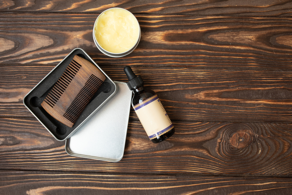 Beard oil on a wooden table alongside a wooden comb and a jar of beard balm