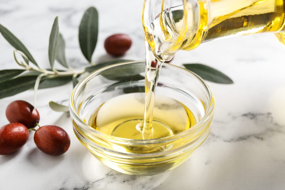 Image of a person pouring jojoba oil for hair into a glass jar with oil seeds on a table next to leaves
