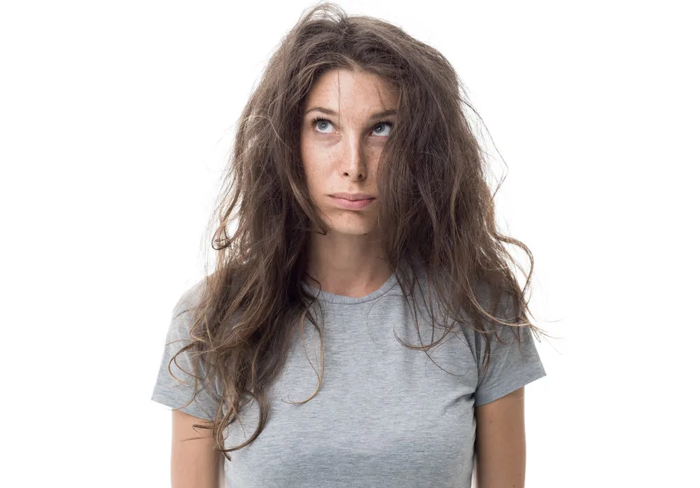 Woman with messy hair looking upward at the top of her head in a grey shirt wondering how to get rid of cowlicks