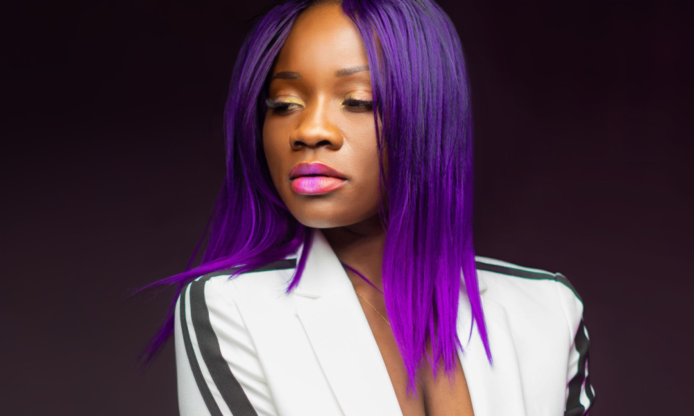 African American woman glances to the side with a white blazer on and dark purple hair with electric purple ombre