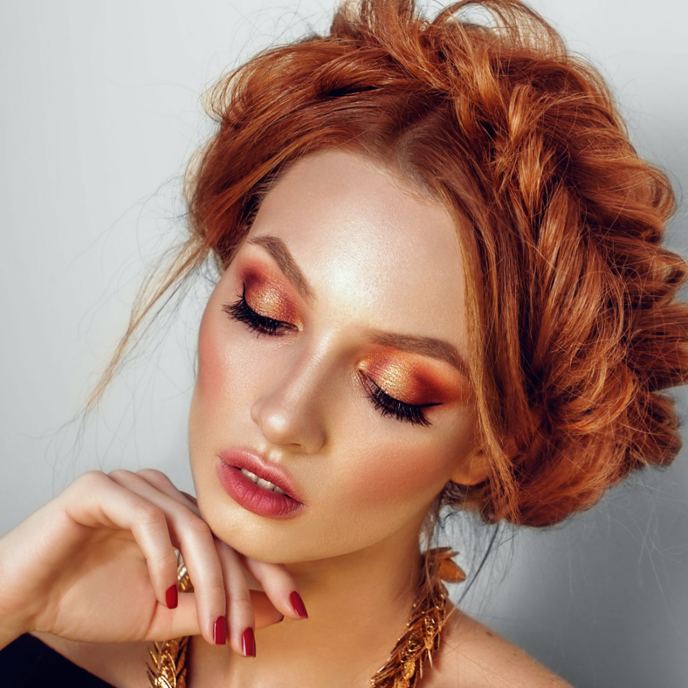 Redheaded woman wearing copper glam makeup look has her hair styled in a fishtail crown braid with hand posed under her chin