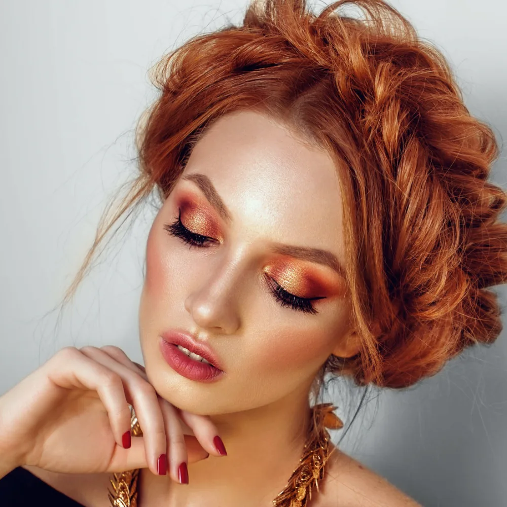 Redheaded woman wearing copper glam makeup look has her hair styled in a fishtail crown braid with hand posed under her chin