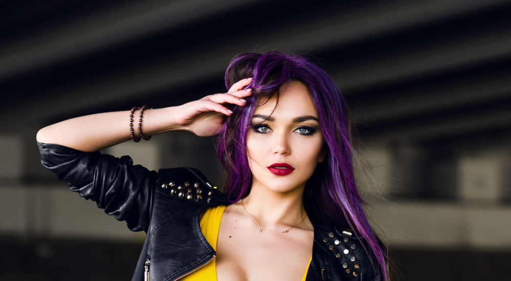 Woman poses with her hand on her head in leather jacket with studs showing off a dark purple hair color
