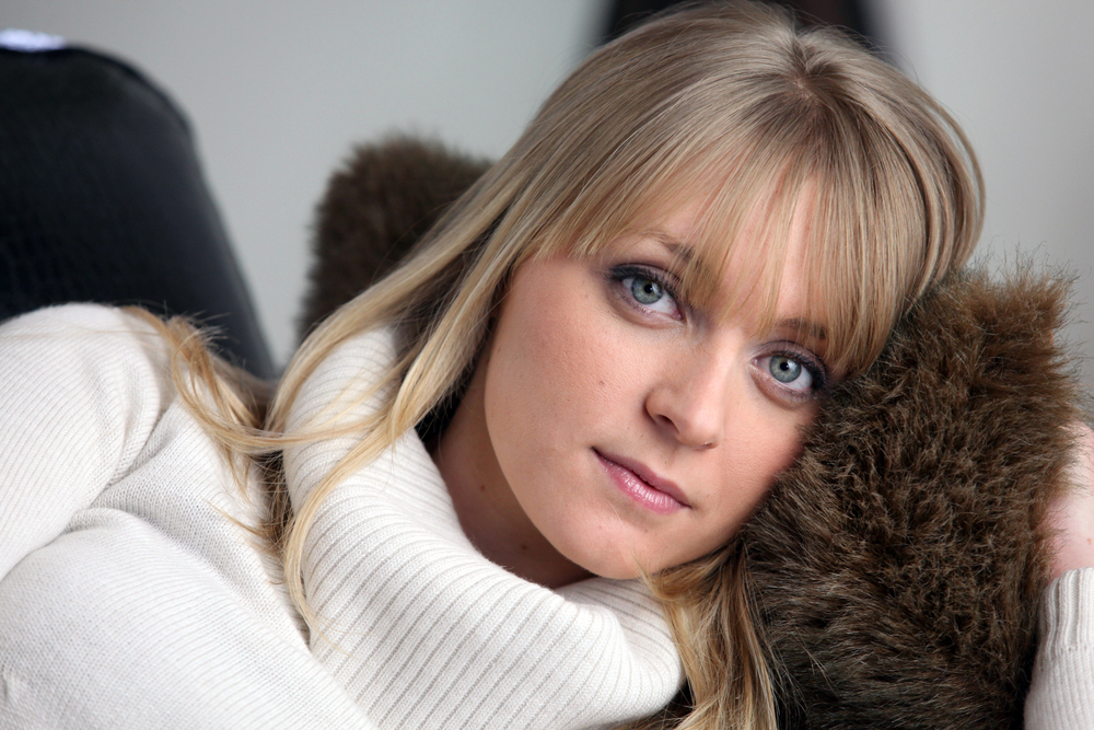 Woman lying down in white turtleneck sweater looks at camera with blonde wispy bangs and straight hair